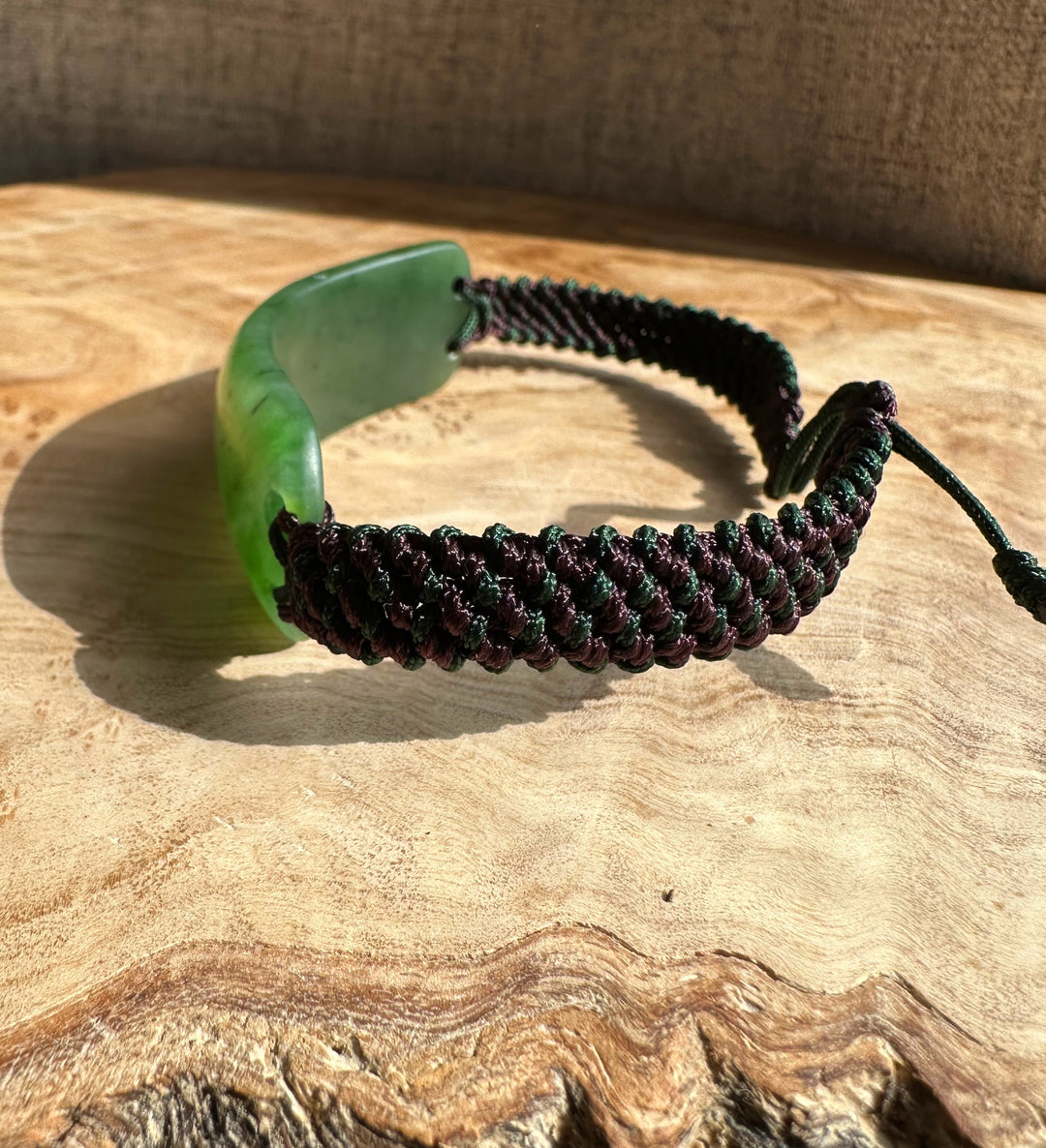 Matte finish bracelet with a curved green jade centerpiece and a maroon and green braided cord on wood.