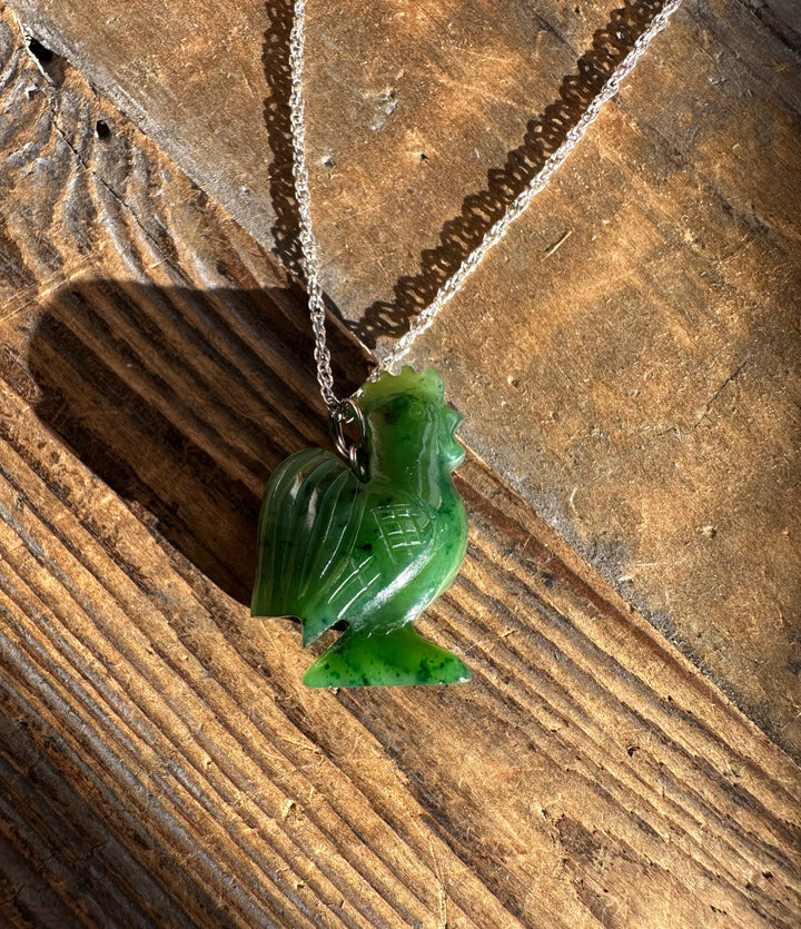 Green jade rooster pendant on a silver chain laid across rough wooden surface