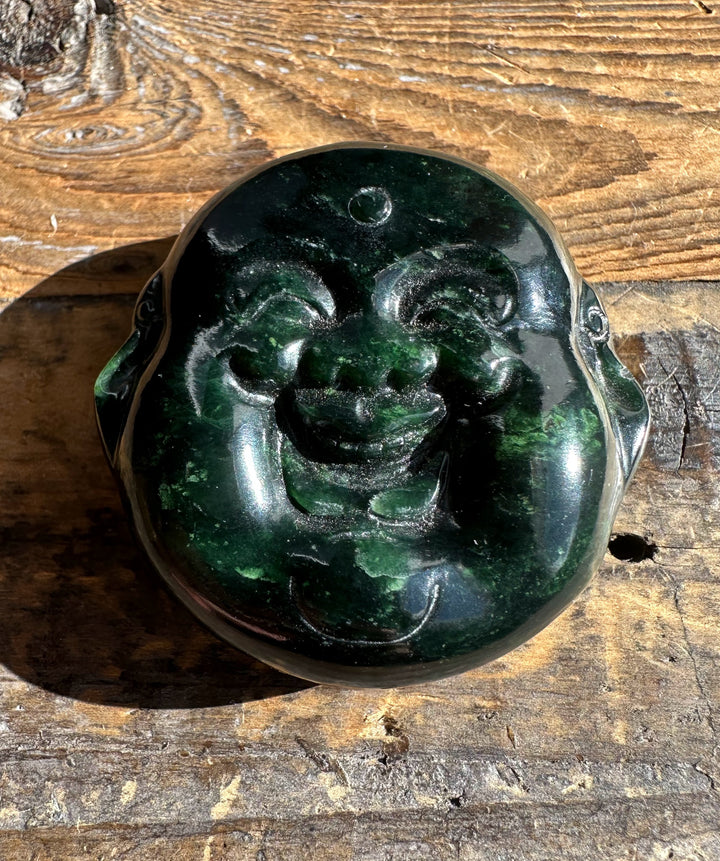 Dark midnight green Canadian jade double sided Happy Buddha head pendant polished on a wooden surface
