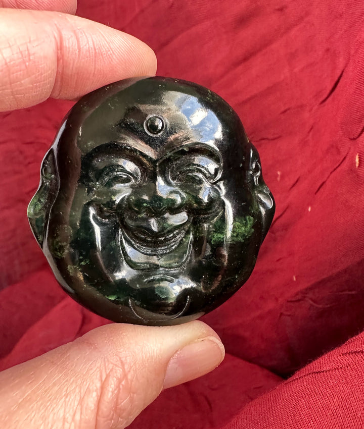 Hand holding a 50mm dark midnight green jade Happy Buddha head against a red fabric background