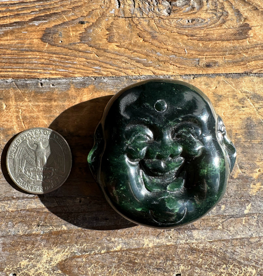 Dark Midnight Green Canadian Jade double sided Happy Buddha head beside a quarter on a weathered wooden surface