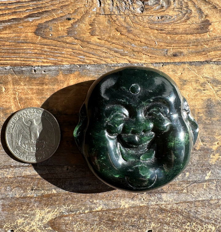 Dark Midnight Green Canadian Jade double sided Happy Buddha head beside a quarter on a weathered wooden surface