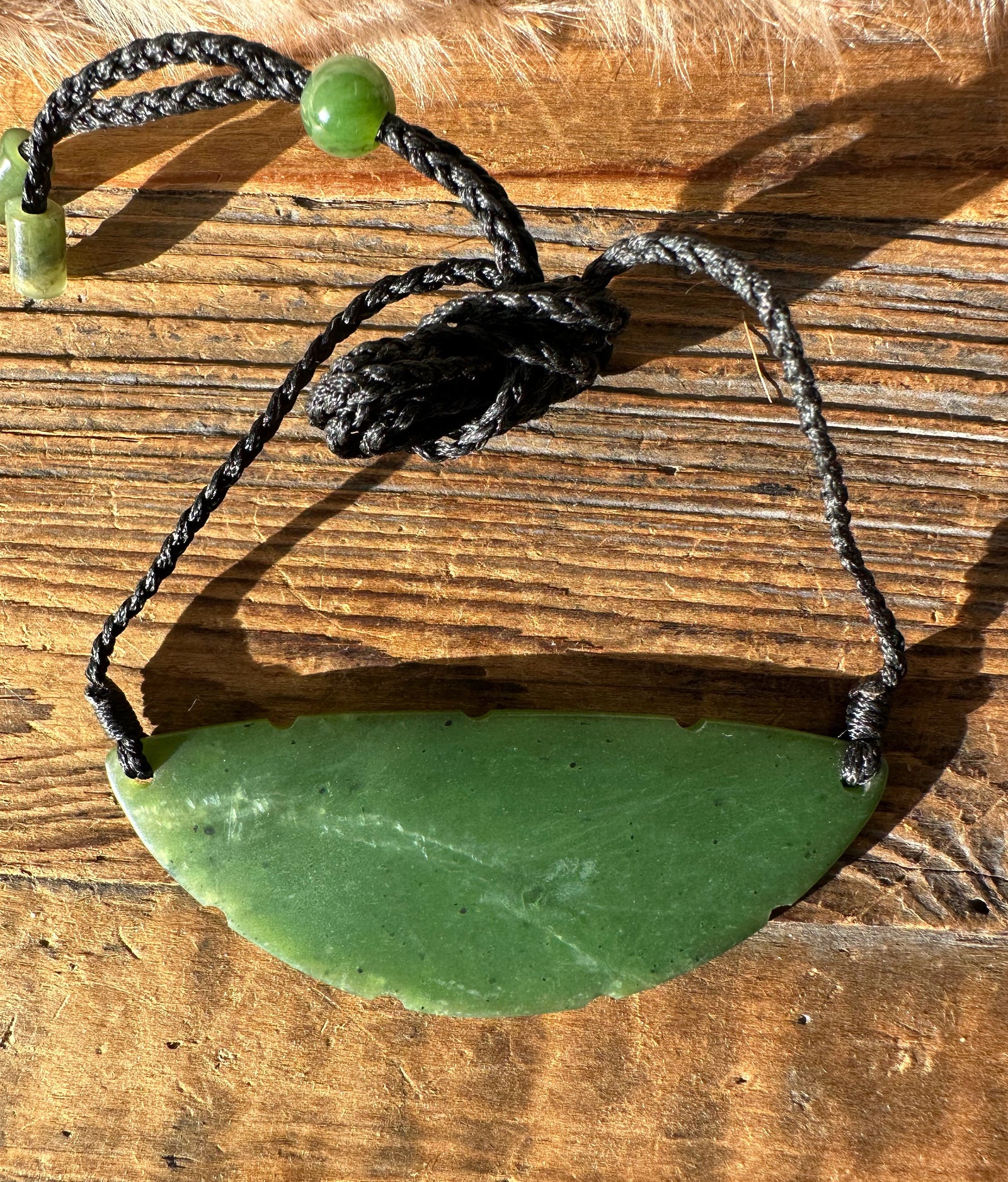 Green jade crescent pendant on a black braided cord with a green bead and knot on a wooden surface