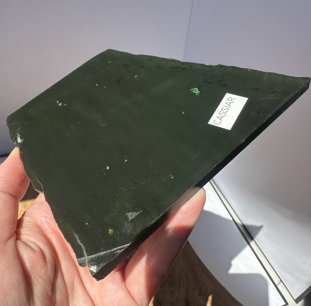 A hand holds a polished dark green stone slab with tiny green inclusions and a small white label reading &