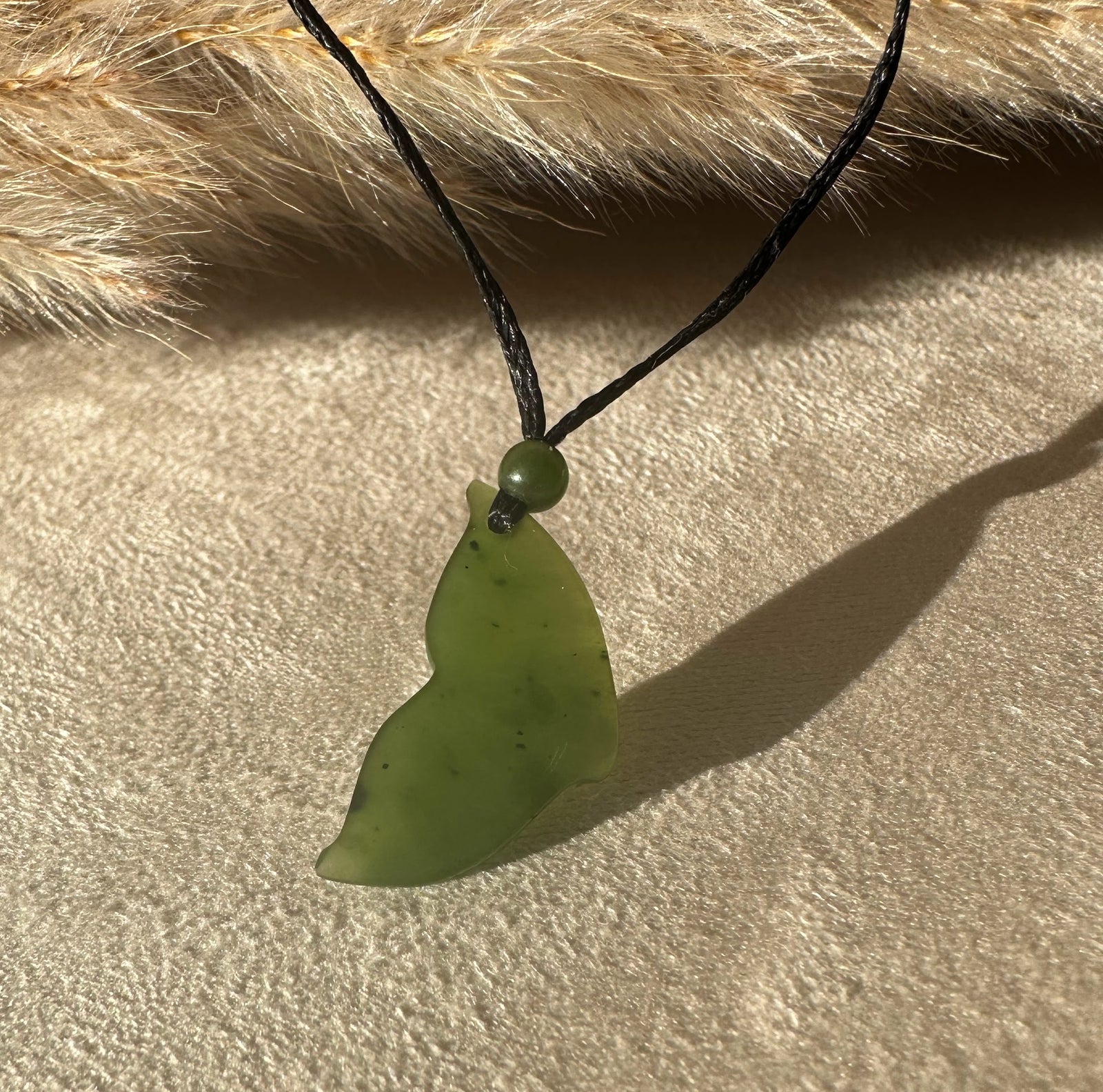 Green jade whale tail pendant on a black cord against a sandy beige background.