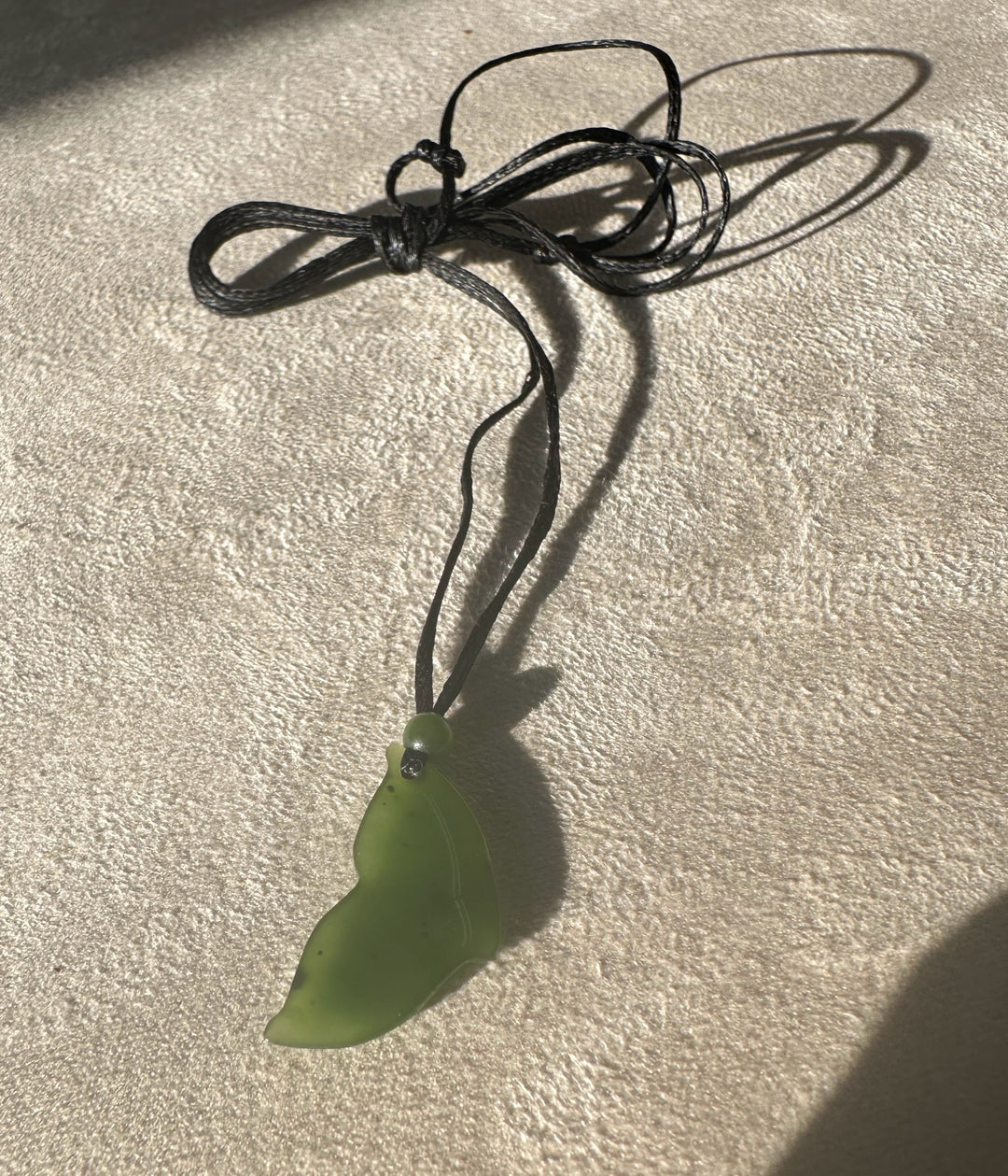 Green jade whale tail pendant on a multi loop black cord resting on a textured beige surface.