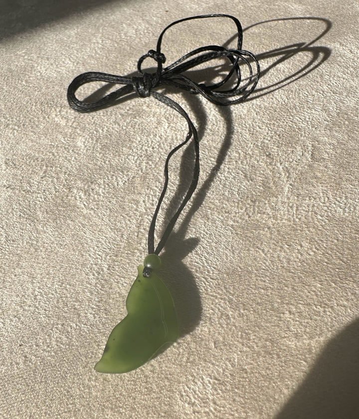 Green jade whale tail pendant on a multi loop black cord resting on a textured beige surface.