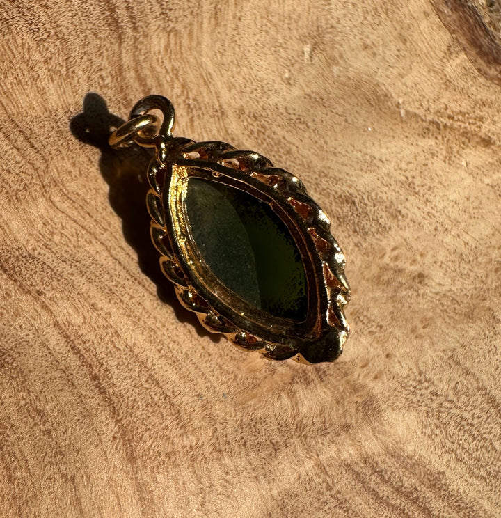 Dark green jade marquise pendant with a gold scalloped frame resting on a rustic wooden surface.
