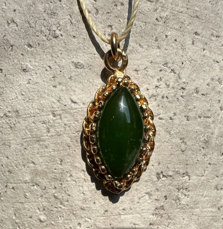 Close up of jade marquise pendant with a gold ornate frame on a textured stone surface.