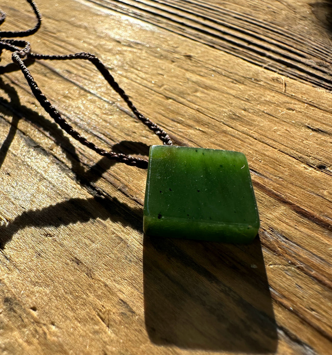 Canadian Jade Square Pendant 15mm resting on a sunlit weathered wooden surface with brown cord.