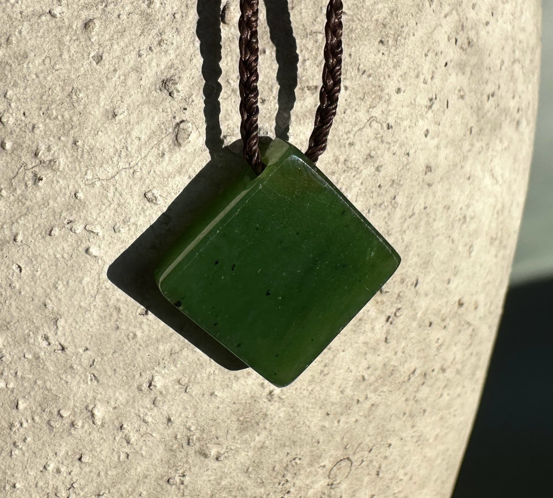Green jade square pendant on a brown braided cord against a light textured background