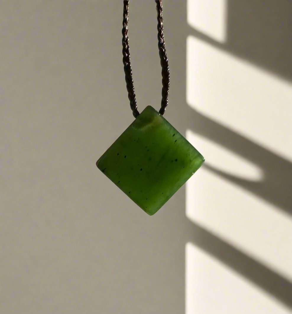 Canadian Jade Square Pendant 15mm suspended from a dark braided cord against a sunlit pale wall.