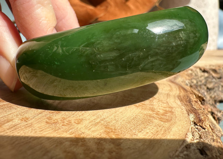 Dark green jade oval bangle 52.5mm held between fingers over a wooden surface.