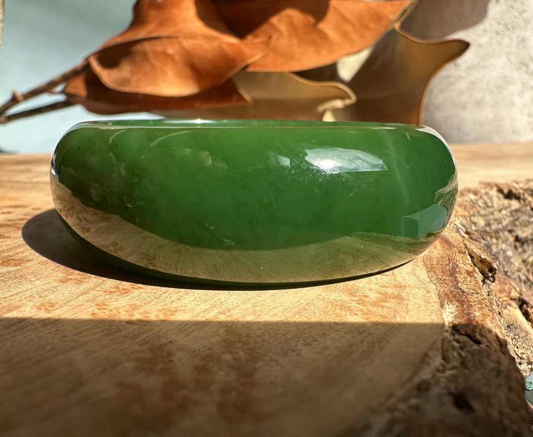 A+ Canadian Jade Oval Bangle in rich green rests on a wooden surface with a dried leaf in the background.