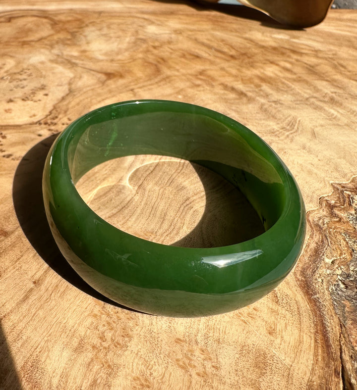A+ Canadian Jade Oval Bangle 52.5mm in vibrant green on a wooden tabletop with a polished glossy finish.