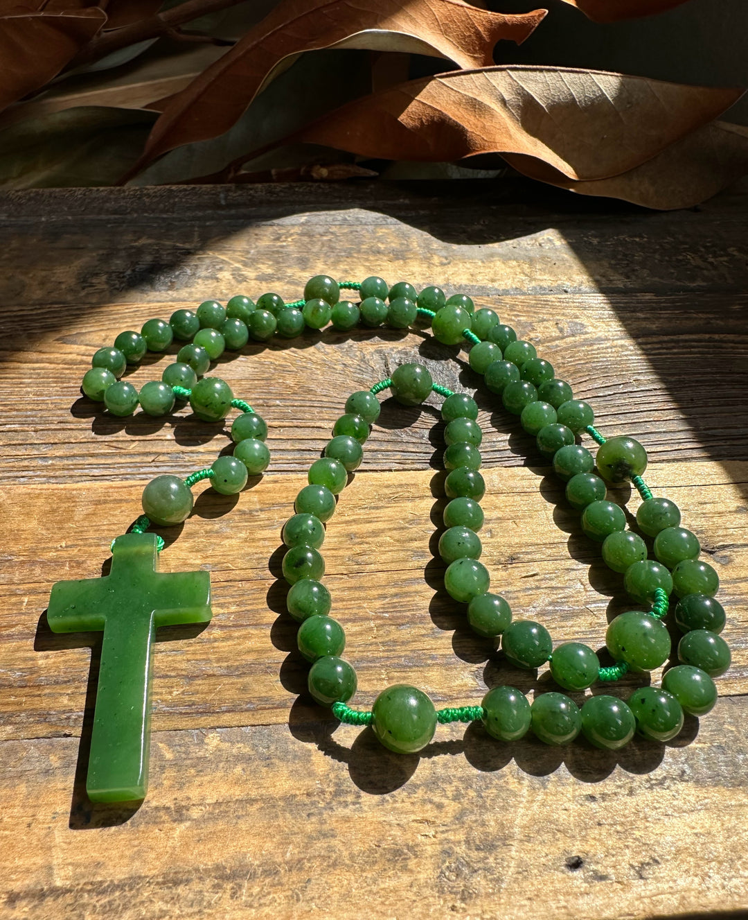 Jade bead rosary with green cross pendant resting on wood beside dried leaves