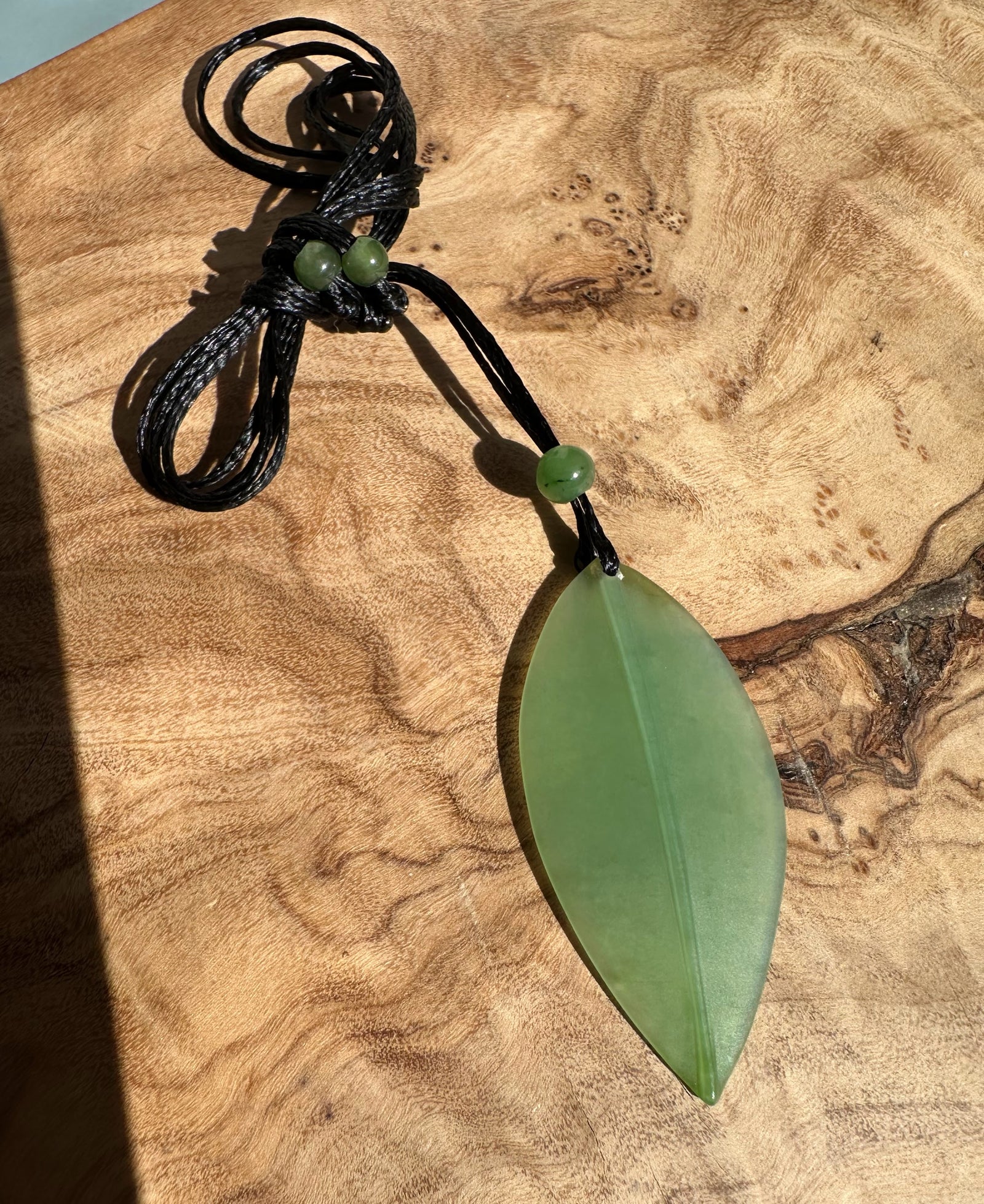 Siberian Jade Leaf Pendant 42mm on a black cord with green beads, placed on a wooden surface.