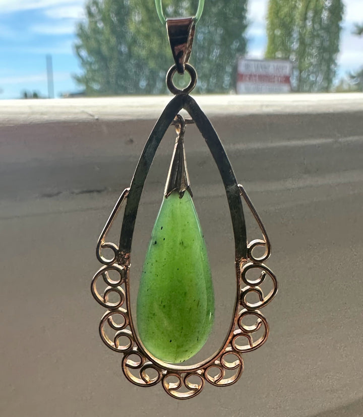 Vintage Canadian nephrite jade teardrop pendant hanging from a metal loop with a blurred outdoor backdrop.