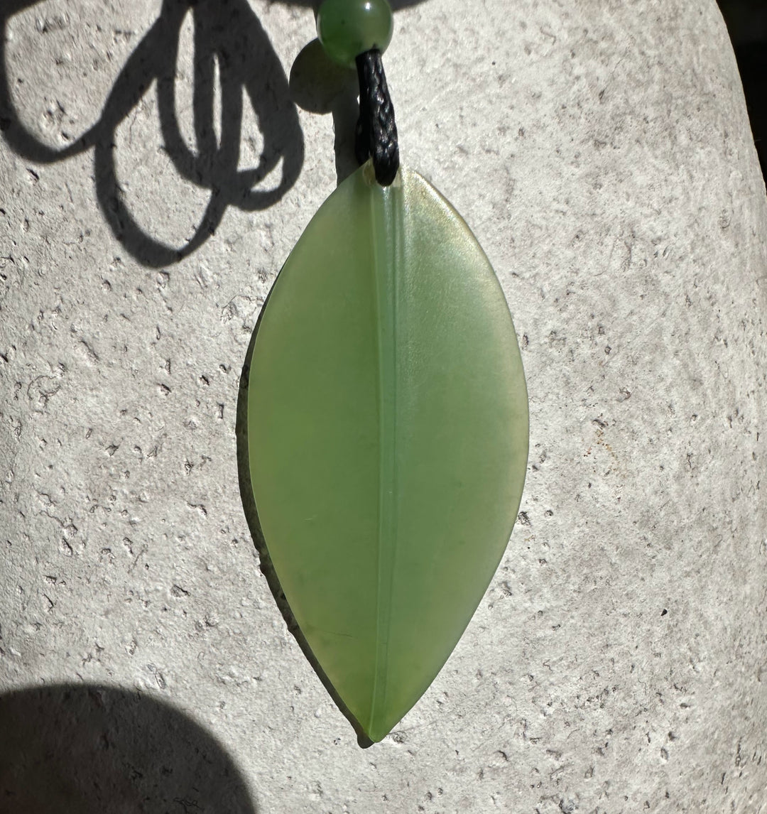 Close-up of pale green jade leaf pendant on a black cord with a bead, against a light stone background.