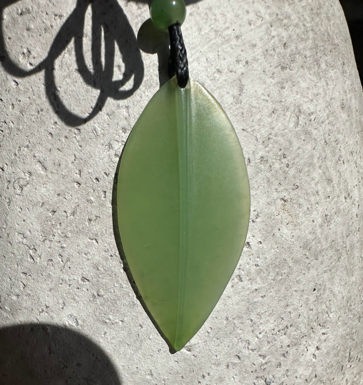 Close-up of pale green jade leaf pendant on a black cord with a bead, against a light stone background.