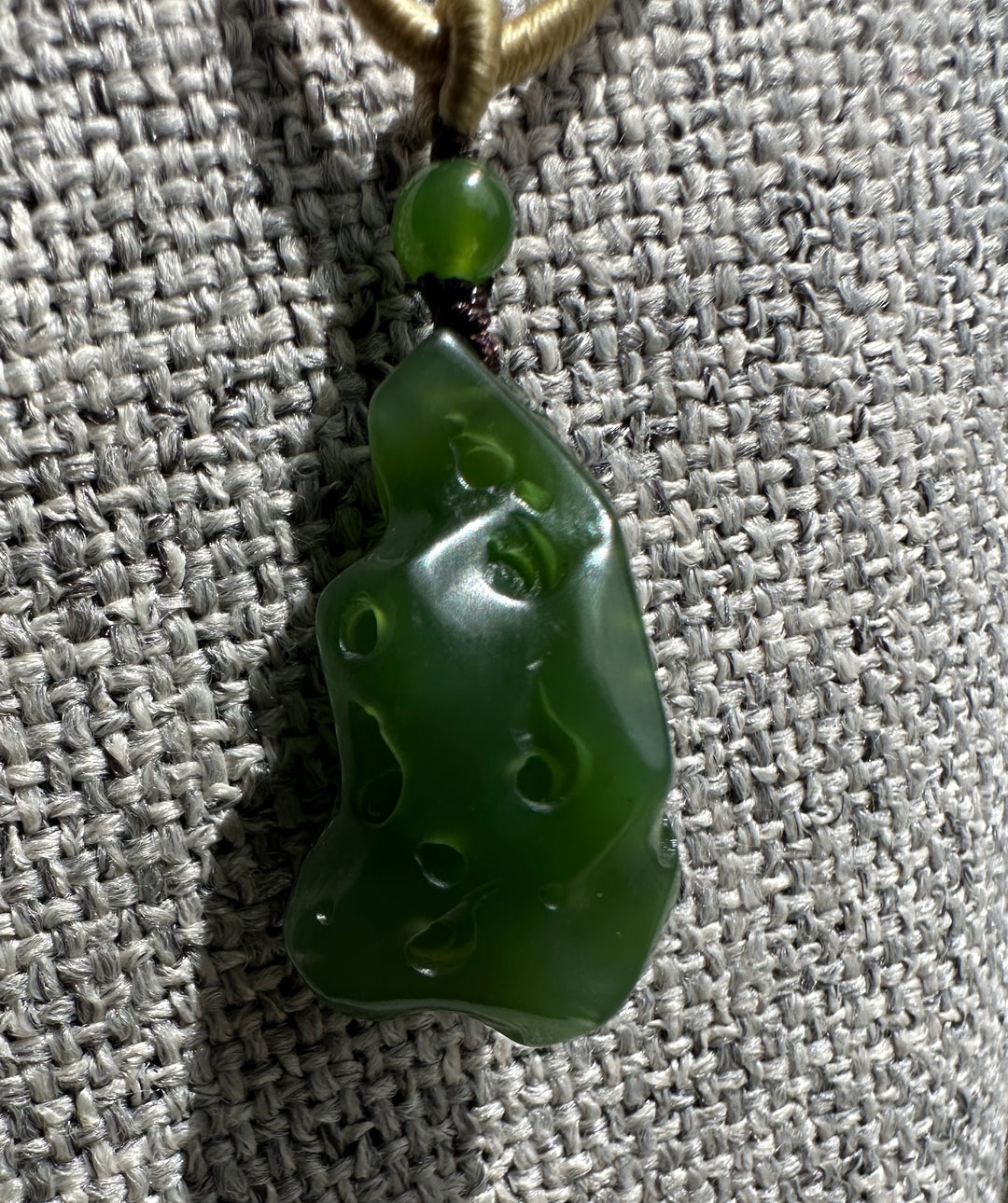Glossy green jade pendant with circular holes against a gray woven fabric background