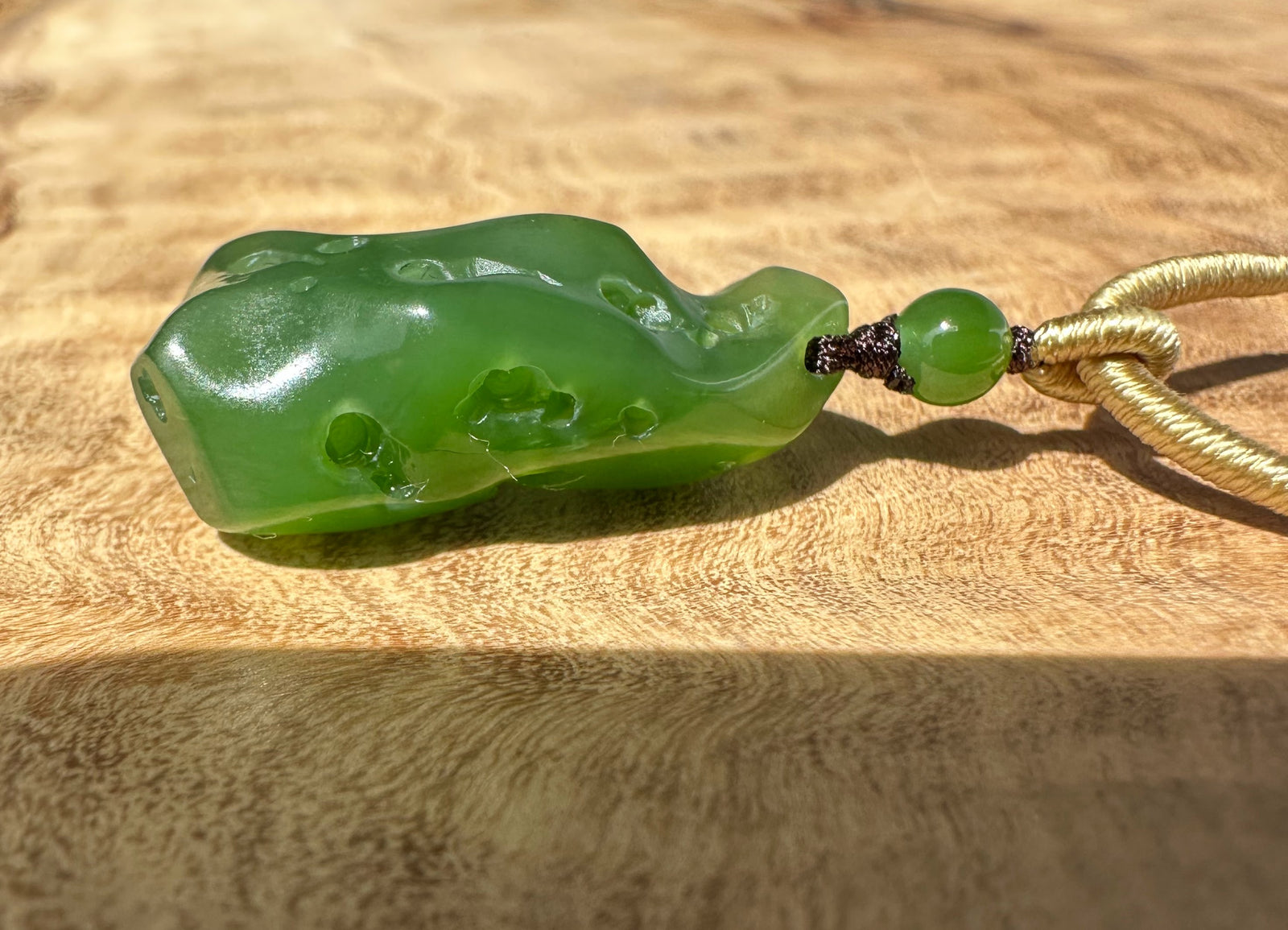 Close-up of a rich green Siberian abstract pendant with carved holes, tied to a knotted beige cord against textured fabric.