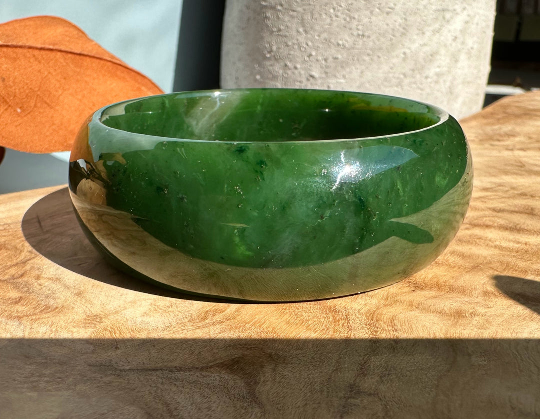 Glossy jade oval bangle 68mm in deep Canadian nephrite jade resting on a wooden table in sunlight.