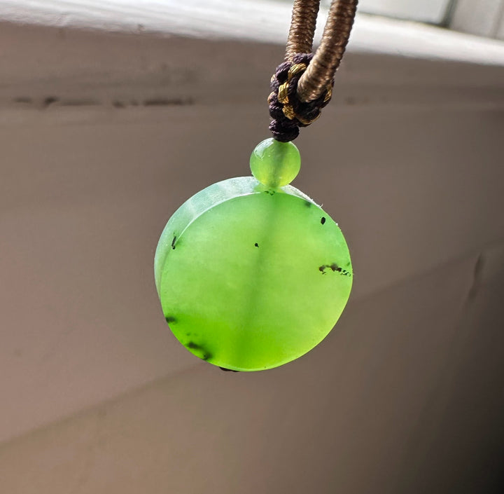 Apple green jade pendant with a round disk shape hanging from a braided brown cord and decorative knot
