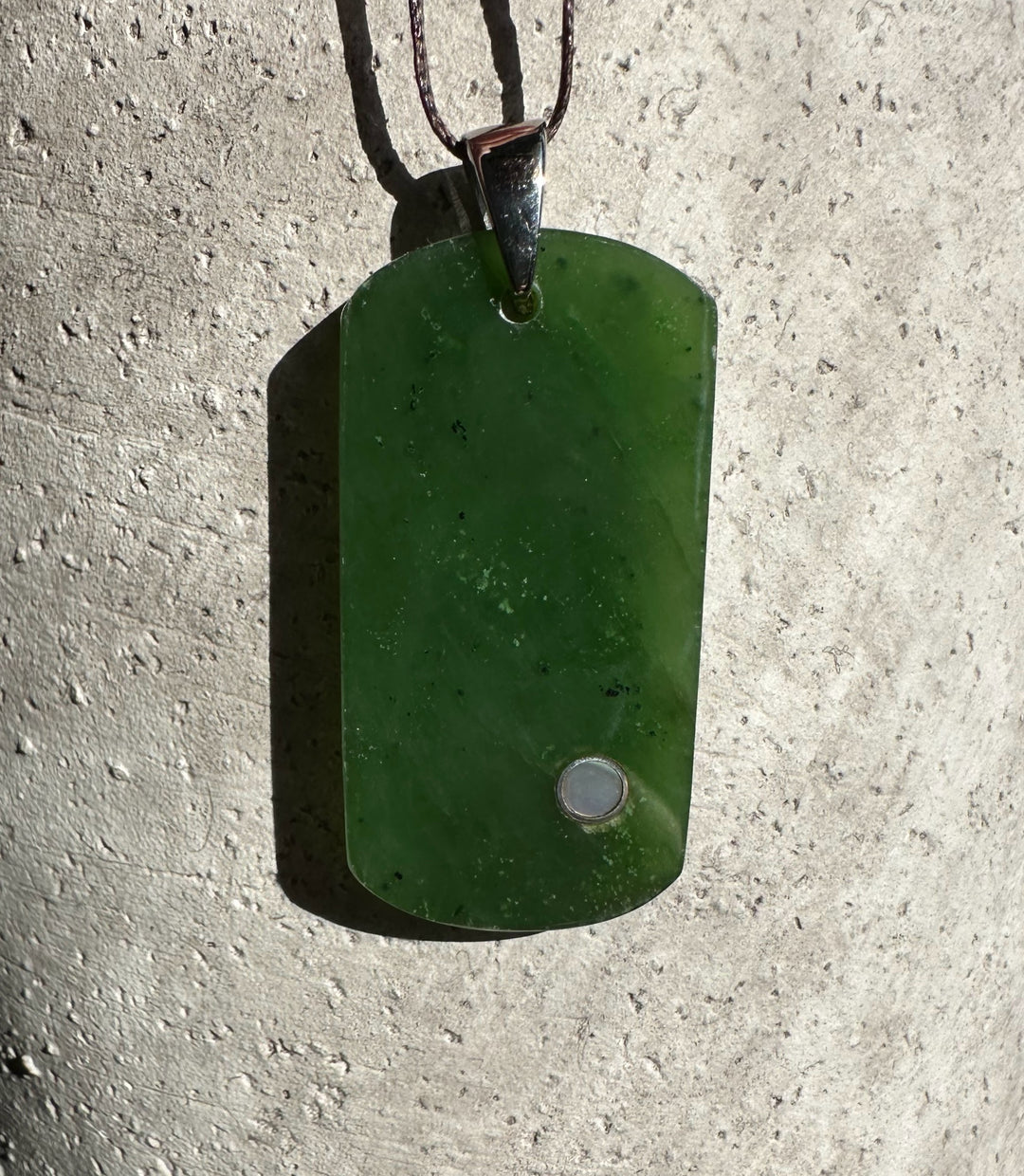 Deep green jade dog tag pendant with a silver bail against a concrete surface