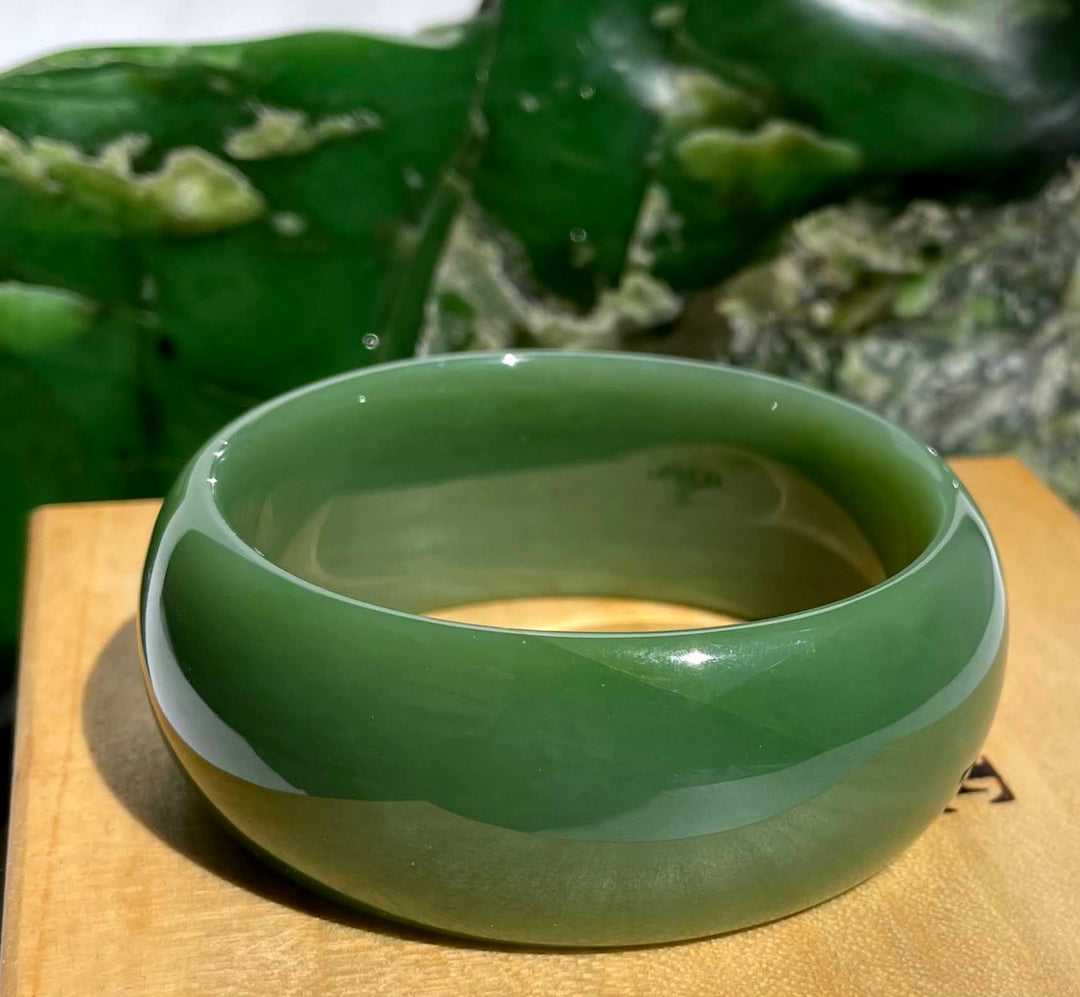 AA Siberian Bangle in deep green jade resting on a wooden surface with a blurred leafy background
