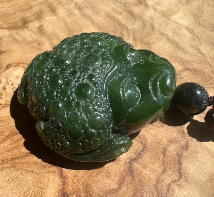 A carved green jade-like frog pendant  with a glossy finish, resting on a textured wooden surface next to a dark bead.