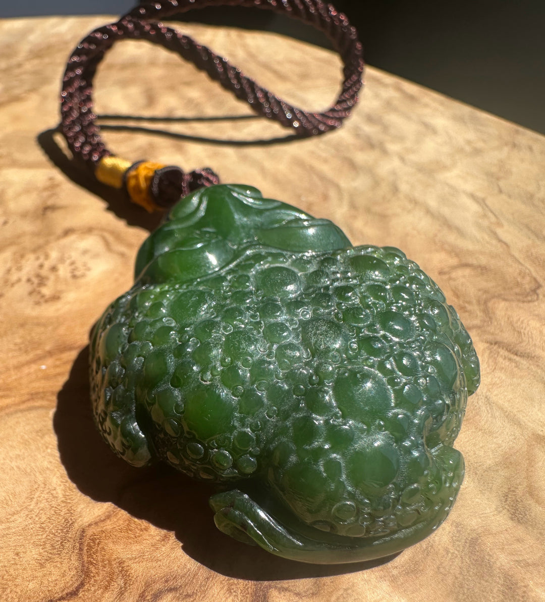 A carved green jade-like frog pendant with a dark brown braided cord and a small yellow accent bead, displayed on a wooden surface.