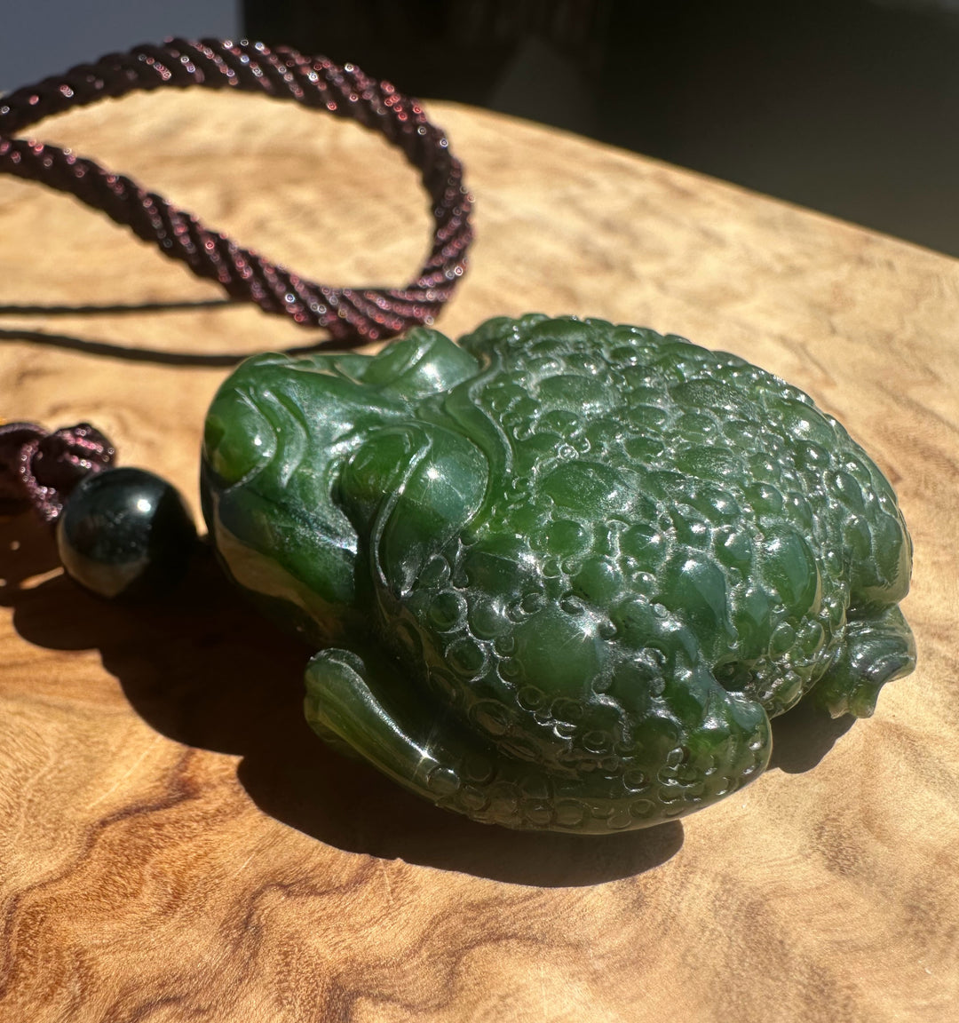 A carved green jade-like frog pendant with a dark brown braided cord and a small dark bead, displayed on a wooden surface.