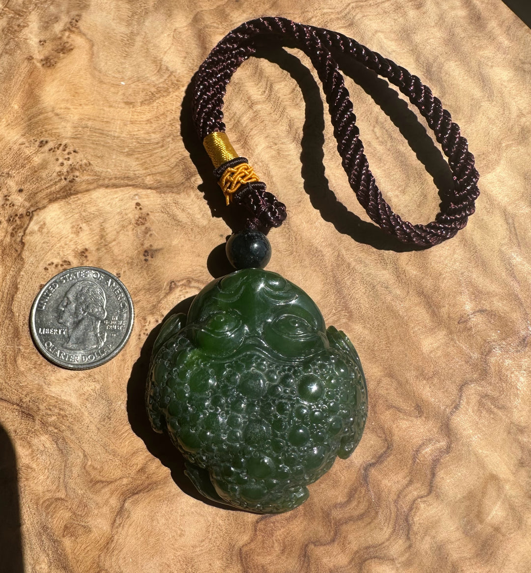 A carved green jade-like pendant with a detailed animal face, hanging from a dark brown braided cord with a small accent bead, displayed on a textured wood surface next to a US quarter for scale.