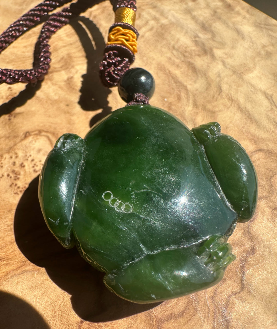 A carved green jade frog pendant necklace with a dark brown and gold braided cord, displayed on a textured wooden surface.