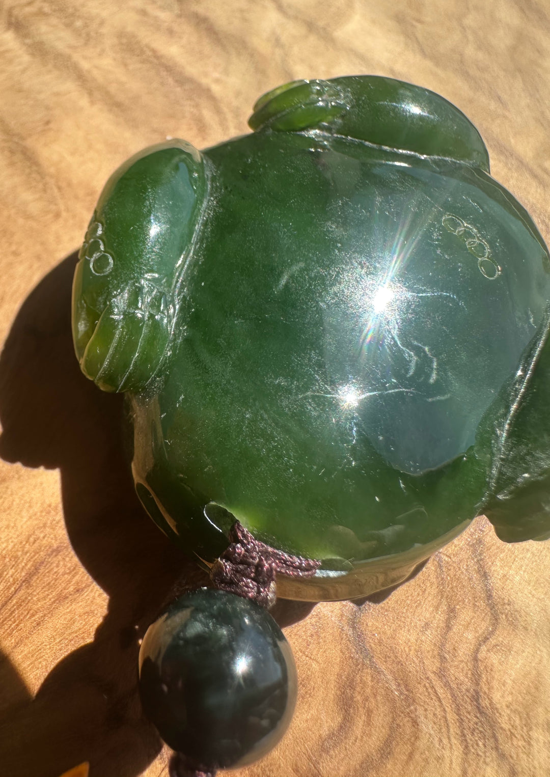 A polished green jade-like frog figurine with a dark hanging bead, photographed close up on a wooden surface.