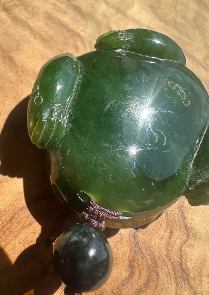 A polished green jade-like frog figurine with a dark hanging bead, photographed close up on a wooden surface.