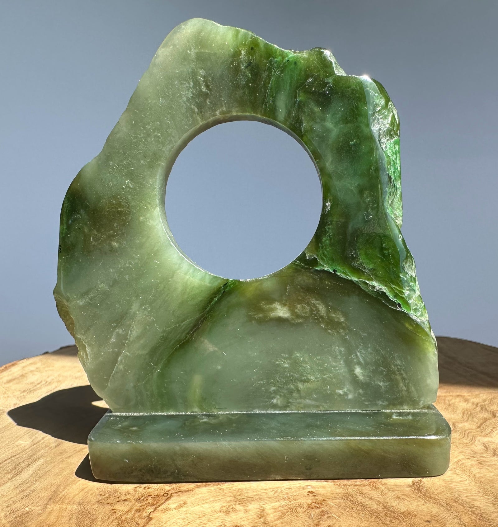 A green jade with a circular hole in the center and a rectangular base, displayed on a wooden surface against a plain gray background.