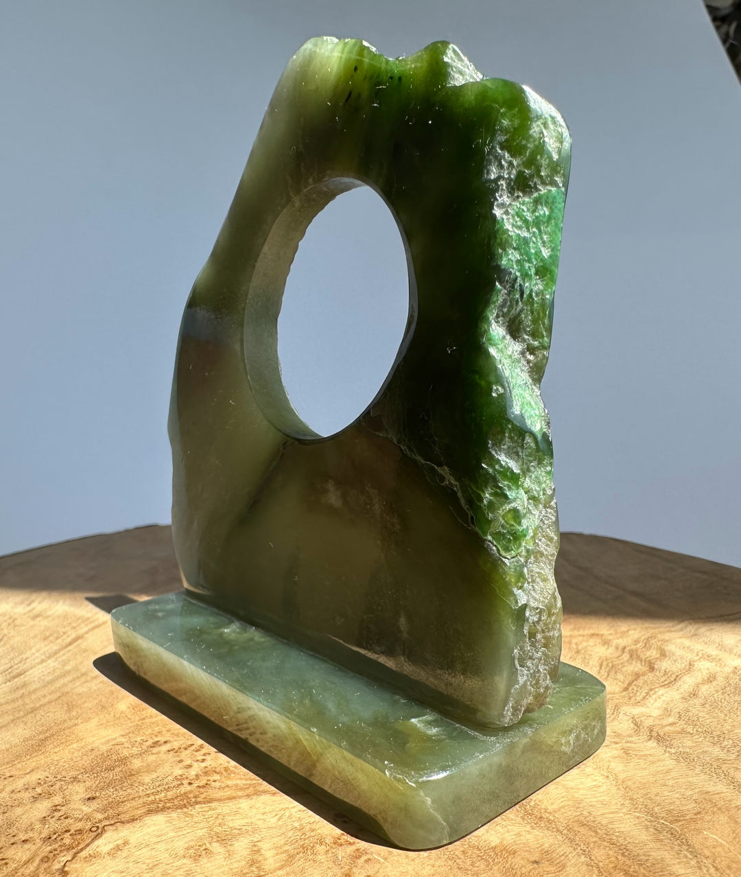 A carved green jade with a circular hole and a rectangular base, displayed on a wooden surface against a light gray background.