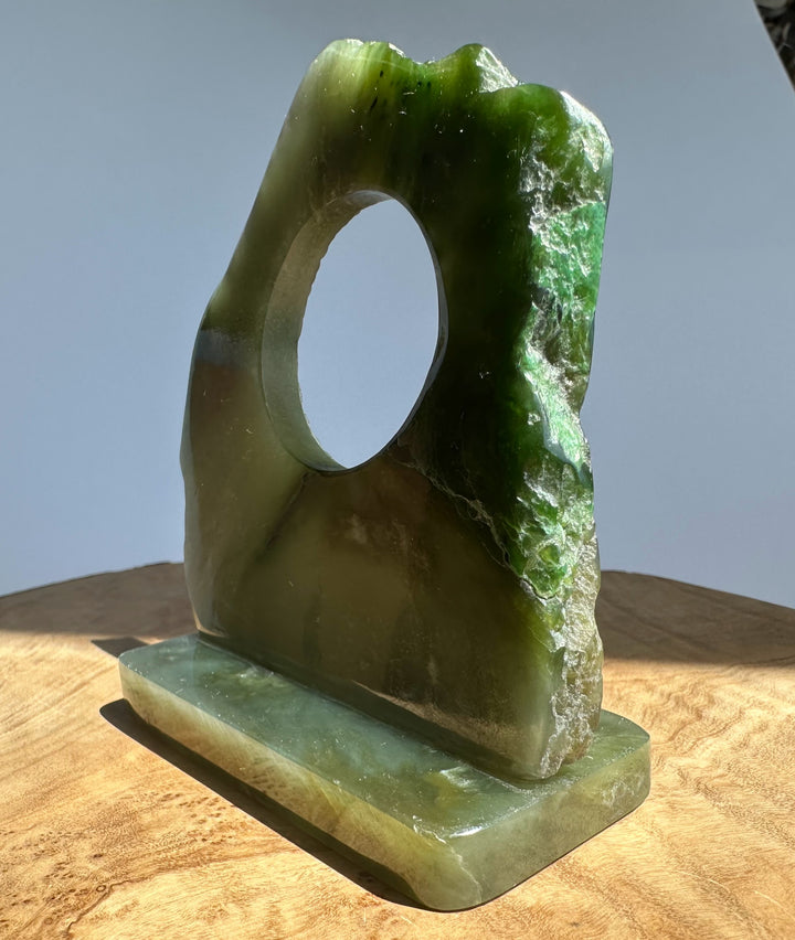 A carved green jade with a circular hole and a rectangular base, displayed on a wooden surface against a light gray background.