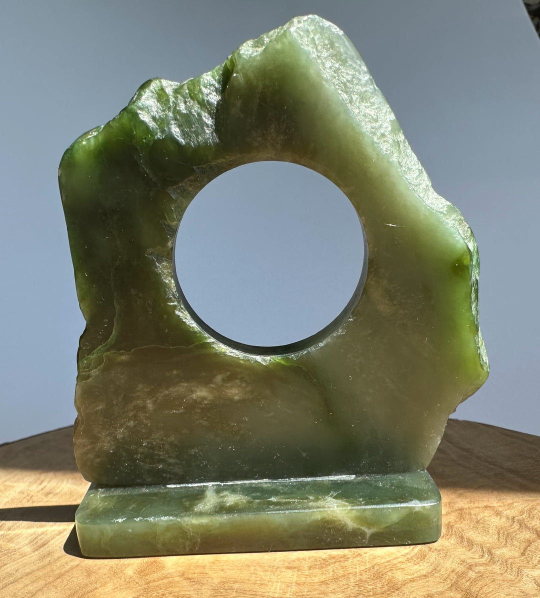 A green jade decorative object with a circular opening and a rectangular base, photographed standing on a wooden surface against a plain light gray background.