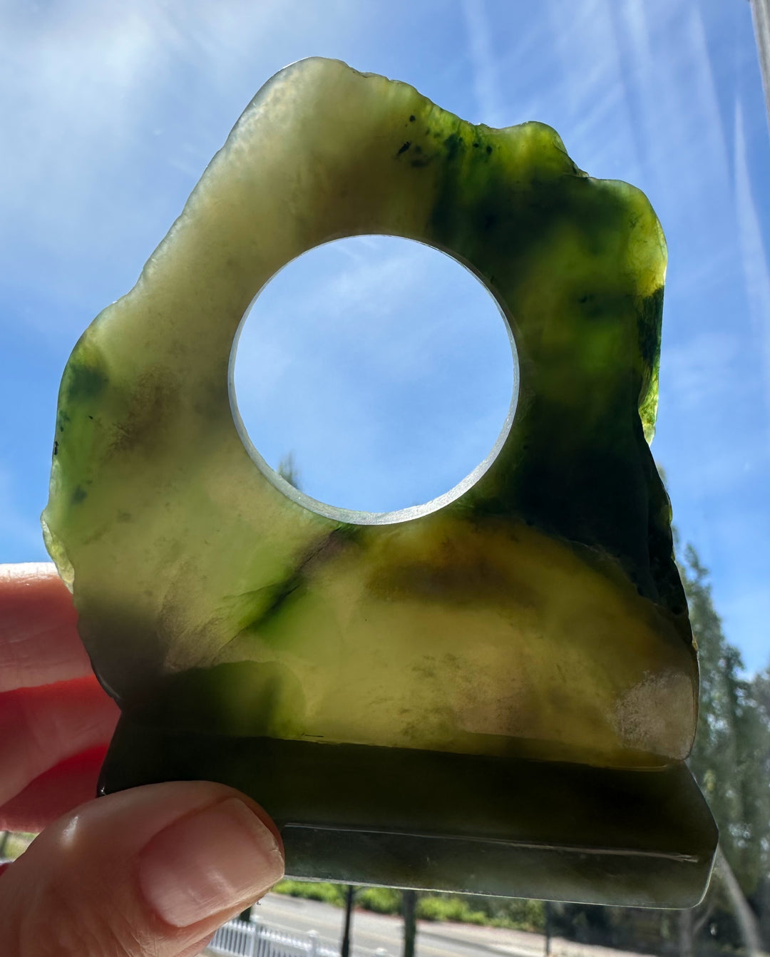 A hand holding a raw, irregular-shaped translucent jade holder against a bright blue sky background.