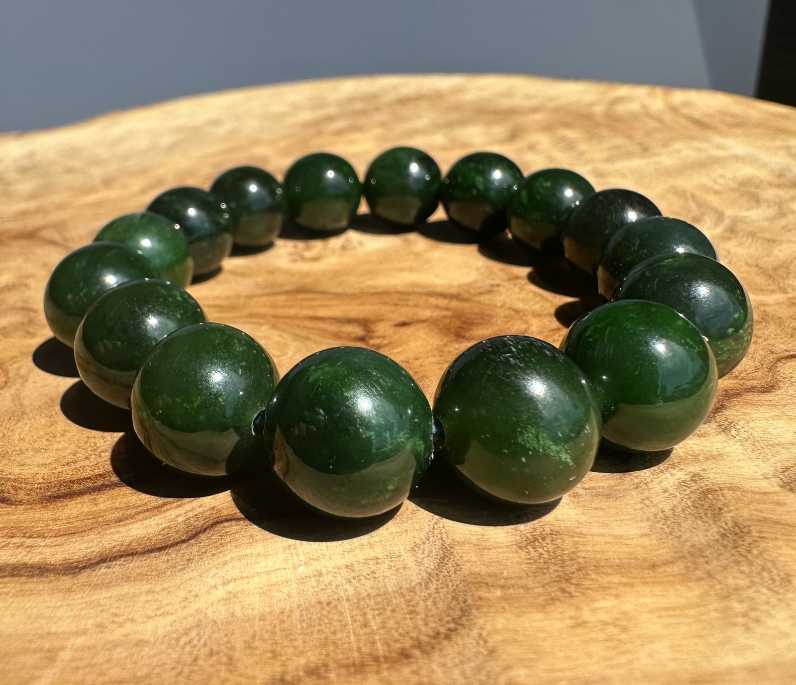 A beaded bracelet made of glossy dark green polished jade beads, arranged in a circle on a natural wood surface.