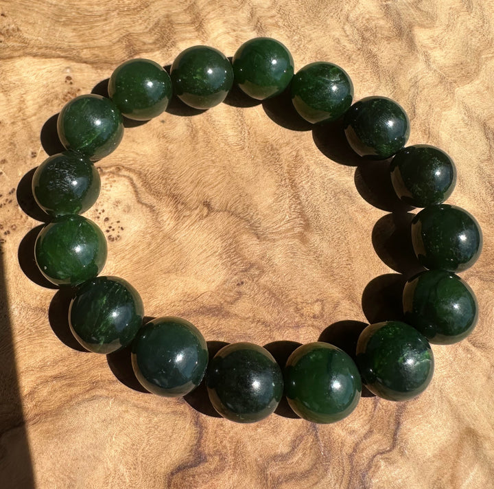 A beaded bracelet made of polished dark green jade round beads, arranged in a circle on a textured light brown wood surface.