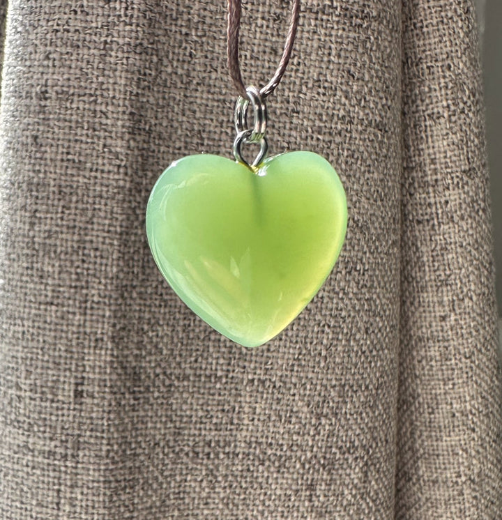 Green Siberian jade heart pendant hanging from a brown cord against textured fabric
