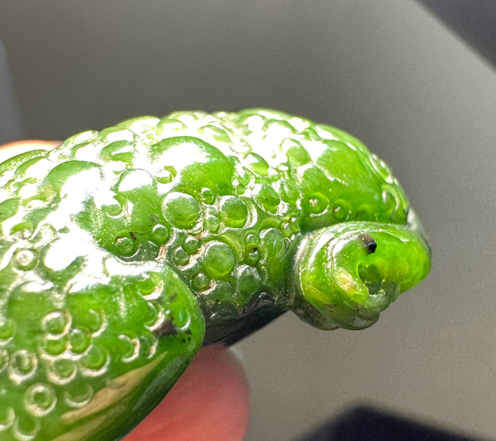 A close-up of a glossy green carved figurine or decorative object with a textured, bubble-like surface and a small protruding eye detail, held against a blurred gray background.