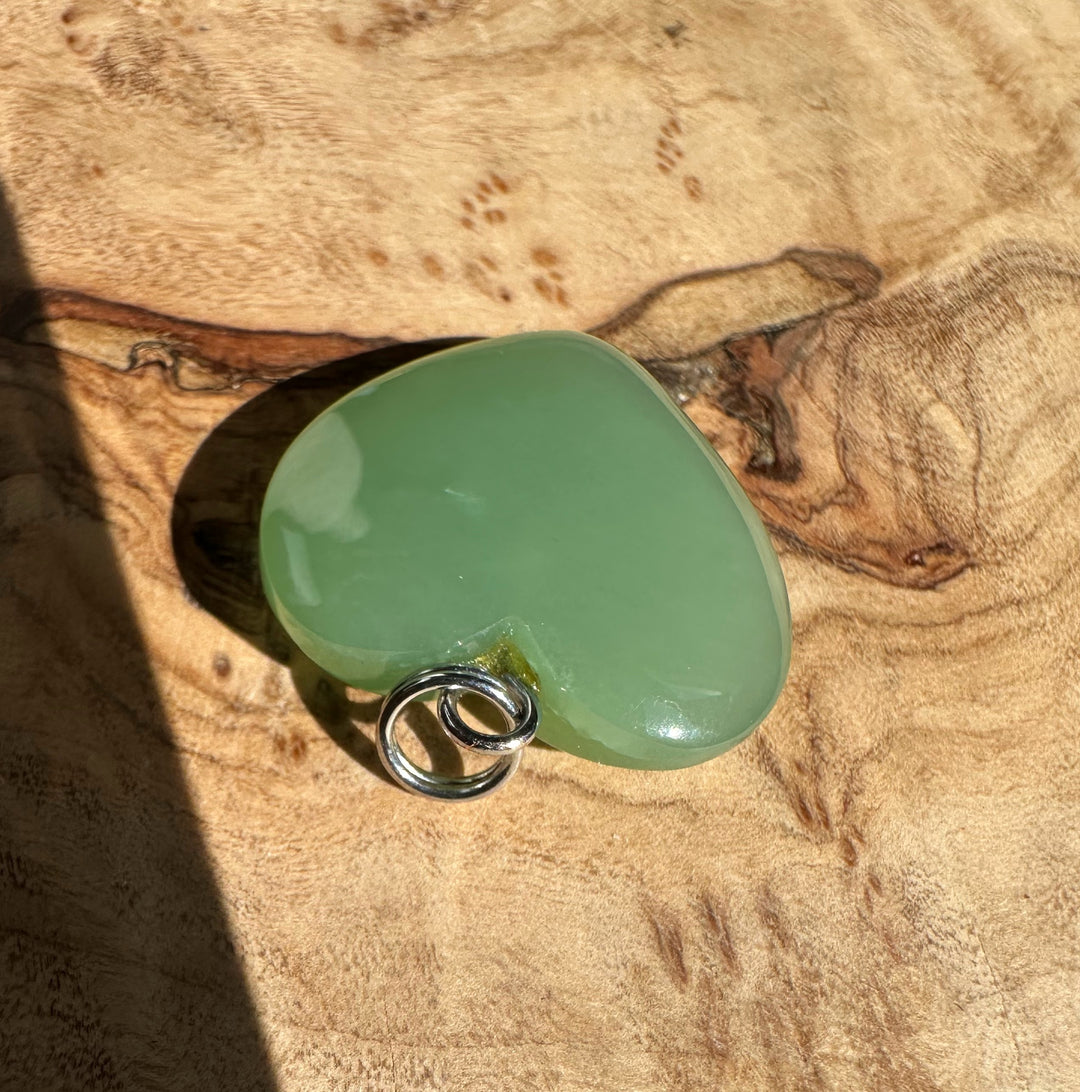 Siberian jade heart pendant in pale green with a silver loop bail on a wooden surface