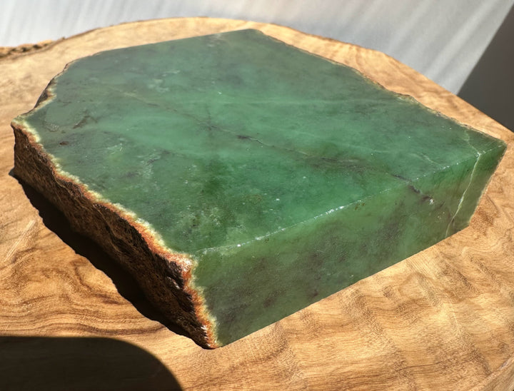 A green nephrite stone block with a rough natural edge, displayed on a rustic wood slice.