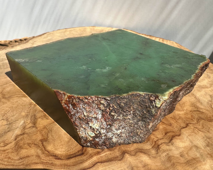 A raw green stone slab with a rough, textured brown and beige edge, displayed on a natural wood slice against a light gray background.