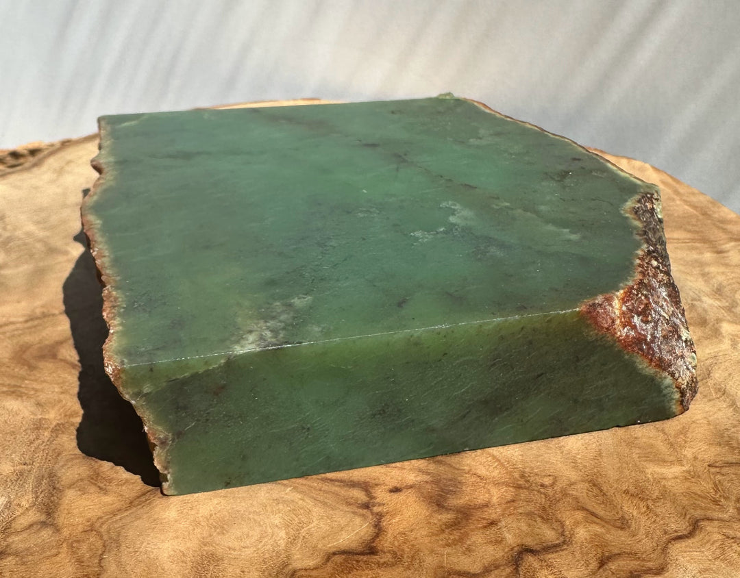 Green jade rectangular slab with rough natural edges, displayed on a curved wood slice against a light neutral background.
